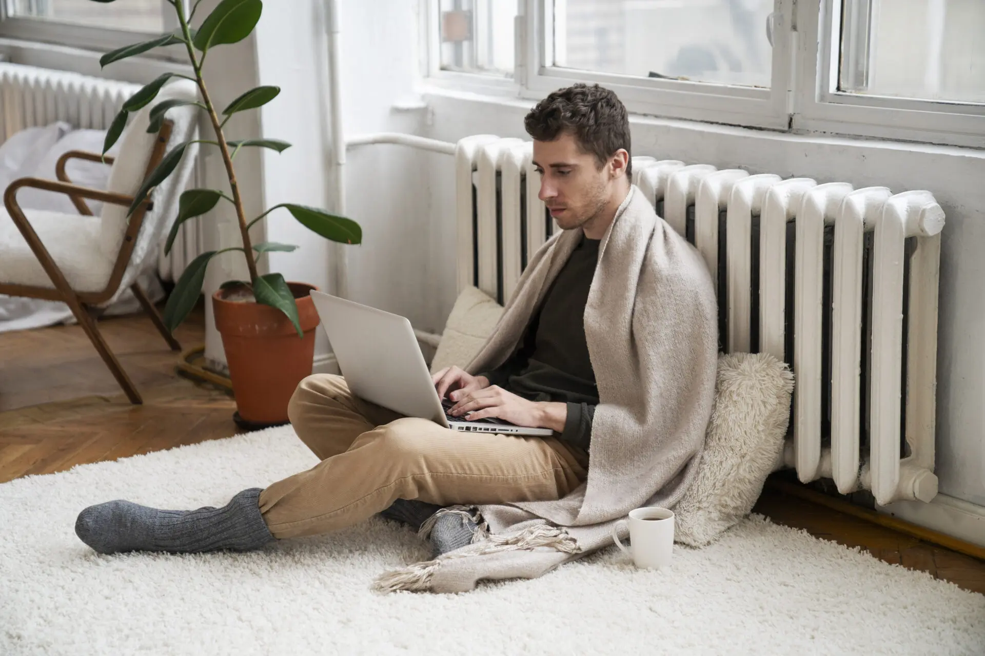 muž zakrytý dekou, sedí pri radiátore, na kolenách má notebook